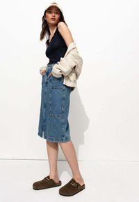 Jupe midi en denim avec deux grandes poches avant. Débardeur bleu foncé, pull beige oversize et sabots en suède marron avec des clous. Fond blanc.