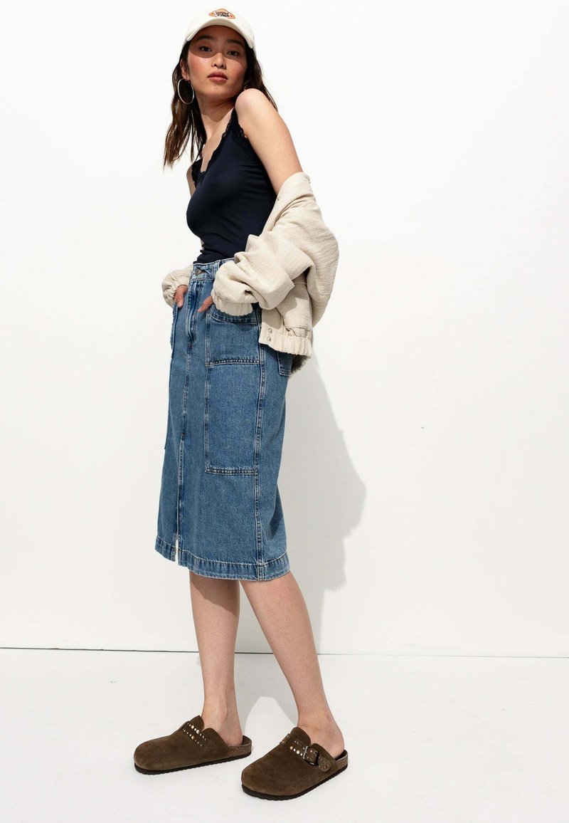 Jupe midi en denim avec deux grandes poches avant. Débardeur bleu foncé, pull beige oversize et sabots en suède marron avec des clous. Fond blanc.