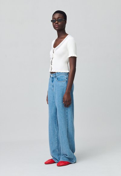 Top bianco a coste con maniche corte e bottoni sul davanti; jeans blu a vita alta e gamba larga; scarpe rosse basse; il modello indossa occhiali da sole scuri.