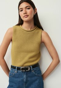 Ung kvinde med langt mørkt hår iført ermeløs sennepsfarvet strikket top og højtaljede blå jeans med sort bælte, stående mod en hvid væg.