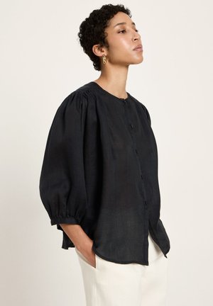 Frau mit kurzen lockigen Haaren trägt eine lockere schwarze Bluse mit Puffärmeln und weißen Hosen, steht mit den Händen in den Taschen.