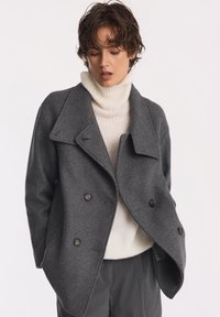 Szary wełniany płaszcz PEACOAT z podwójnym zapięciem na guziki i szerokim kołnierzem, noszony na beżowym golfie, w połączeniu z ciemnymi spodniami.