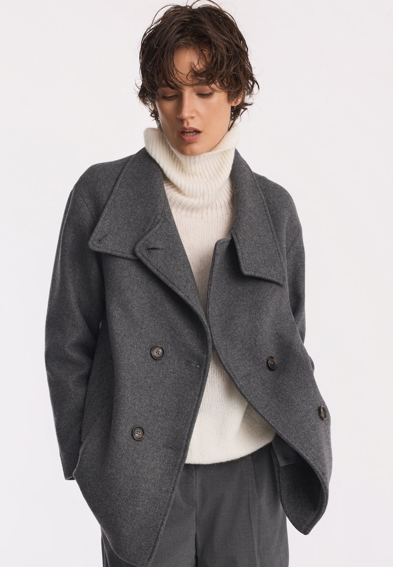 Szary wełniany płaszcz PEACOAT z podwójnym zapięciem na guziki i szerokim kołnierzem, noszony na beżowym golfie, w połączeniu z ciemnymi spodniami.