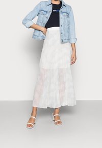 Džínová bunda, námořnický top, průhledná bílá plisovaná sukně a bílé sandály na bloku, představující vrstvený, lehký letní outfit.