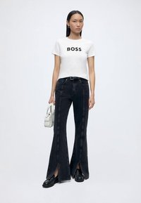 Mujer vistiendo una camiseta blanca con el texto "BOSS", vaqueros negros de cintura alta y pierna ancha con aberturas delanteras, zapatos negros y sosteniendo un bolso blanco.