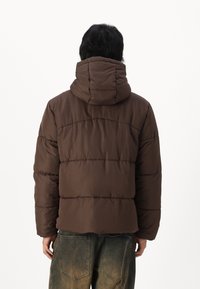 Brun pufjakke med hætte, der har et firkantet design og quiltet tekstur. Jakken har en afslappet pasform og synlige syninger.