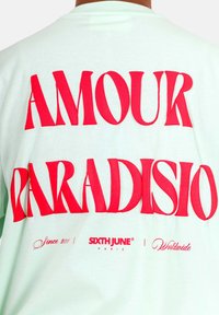 Camiseta verde menta con un atrevido texto gráfico en rojo que dice "AMOUR PARADISIO". Presenta el logo de la marca "SIXTH JUNE" y texto adicional en una fuente más pequeña.