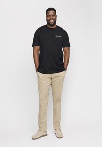 Svart bomullst-skjorte med "Club1990" tekst på brystet, kombinert med beige chinos og lyse sko. Stående posisjon, nøytral bakgrunn.