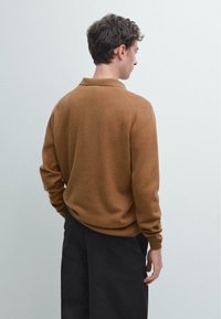 Hnědý pletený pulovr s límečkem, s dlouhými rukávy a žebrovaným lemem, nošený s černými kalhotami. Tkanina vypadá měkká a texturovaná.