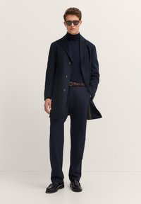 Marineblauer Wollmantel über einem marineblauen Rollkragenpullover, kombiniert mit maßgeschneiderten dunkelgrauen Hosen und einem braunen Gürtel. Die Accessoires umfassen dunkle Sonnenbrillen.