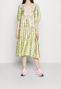Femme portant une robe midi ample à volants avec un imprimé floral vert et violet et des baskets roses et blanches, debout devant un fond uni.