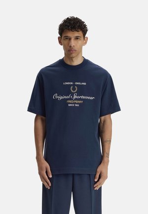 Hombre con cabello corto y rizado que lleva una camiseta azul marino de Fred Perry con texto y gráficos impresos, de pie frente a un fondo liso.