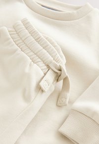 Kermanvärinen collegepaita ja verkkarit, joissa on resorireunukset hihansuissa ja vyötäröllä. Sileä pinta, verkkareissa taskujen kohdalla kerättyä yksityiskohtaa.