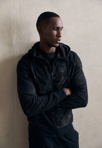 Sudadera con cremallera negra de superficie texturizada, que cuenta con capucha, bolsillos frontales y un corte ajustado, llevada sobre una camiseta negra.