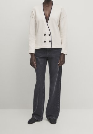 Beige double-breasted jacket with black buttons and trim, paired with dark high-waisted jeans, flat black shoes, and a minimalistic context.