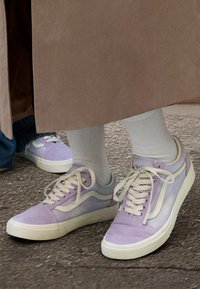 Personne portant des chaussettes blanches et des baskets violet clair debout sur le trottoir, avec une autre personne aux chaussures similaires partiellement visible.