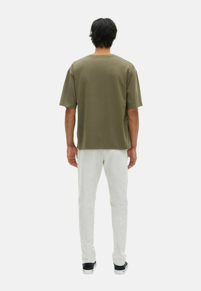 Uomo di spalle che indossa una camicia a maniche corte verde oliva, pantaloni bianchi e sneaker nere su uno sfondo chiaro e uniforme.