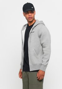 Grijze ritshoodie van zacht materiaal met een voorzak en capuchon. Gecombineerd met een zwart T-shirt en olijfgroene cargobroeken, afgemaakt met een zwarte pet.