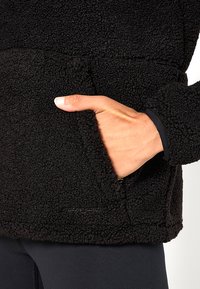 Giacca in pile nera con tessuto testurizzato, dotata di una tasca laterale e dettagli di cucitura visibili. Accentuata con un logo vicino all'orlo.