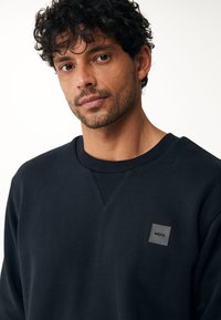 Námořnický crewneck svetr vyrobený z bavlněné směsi. Obsahuje texturovaný límec a malou čtvercovou nášivku loga na hrudi.