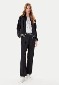 Giacca sportiva nera con zip, abbinata a una t-shirt bianca con grafica. Pantaloni neri con fascia superiore a marchio, indossati con sneakers bianche.