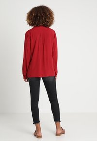 Rood overhemd met lange mouwen en witte stippen, met een geplooide achterkant, gecombineerd met zwarte skinny jeans en beige platte sandalen.