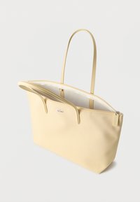 Sac cabas beige clair avec longues poignées, fermeture éclair ouverte et petit logo crocodile sur le devant, sur fond blanc uni.