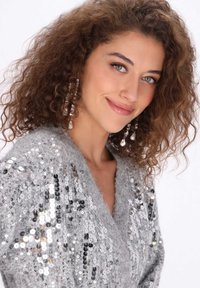 Grijze trui met pailletten, voorzien van een V-hals ontwerp. Heeft een zachte, fuzzy stof en lange, krullende haren met hangende oorbellen.