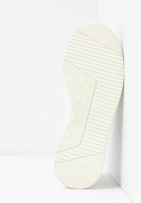 Valkoisen tennarin pohja, jossa on kuvioitu kulutuspinta, keskellä hieman koholla oleva logo ja sileät reunat. Pohjassa on pystysuoria ja vaakasuoria uurteita.