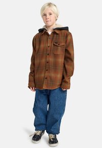 Una camicia di flanella a scacchi marrone e verde con cappuccio nero, chiusura a bottoni e taschino sul petto, abbinata a jeans in denim blu larghi e sneakers nere.