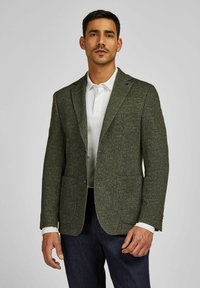 Man in een donkergroene, gestructureerde blazer over een wit poloshirt en donkere spijkerbroek, staand tegen een effen lichtgrijze achtergrond.