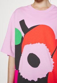 Camiseta rosa con diseños gráficos llamativos en rojo, verde y negro. Corte holgado con mangas cortas y cuello redondo.