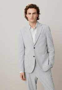 Chaqueta de traje gris claro con tejido texturizado, dos bolsillos frontales, diseño de un solo botón y dos botones. Combinada con una camisa clara debajo.