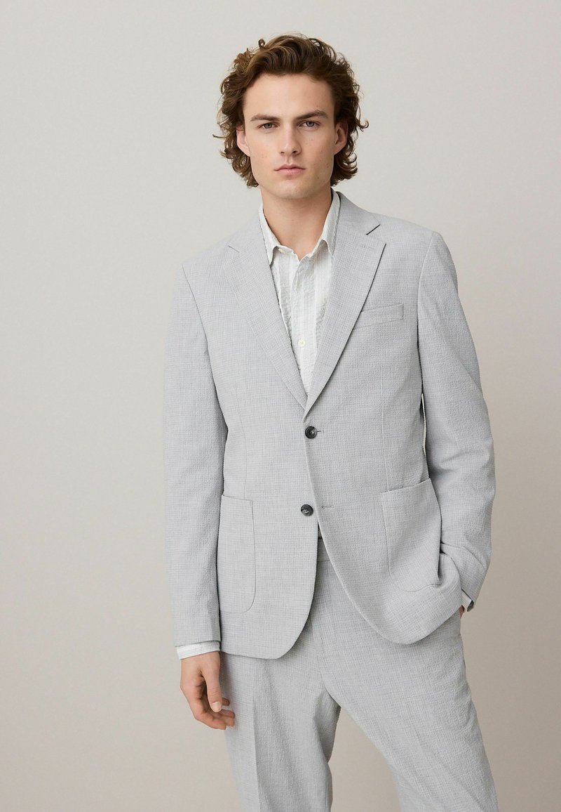Chaqueta de traje gris claro con tejido texturizado, dos bolsillos frontales, diseño de un solo botón y dos botones. Combinada con una camisa clara debajo.