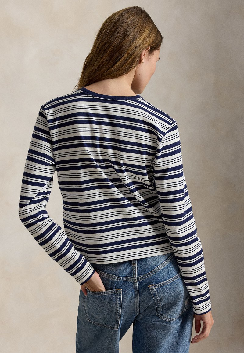 Kvinde med langt hår iført en navy- og hvidstribet langærmet trøje og blå jeans, vendt væk med den ene hånd i baglommen.