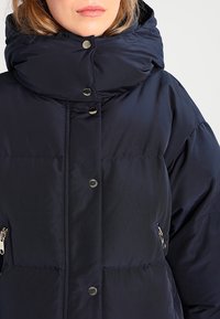 Námořnická pufovací bunda s vysokým límcem, patentkami a dvěma bočními kapsami na zip. Vyrobena z hladkého, izolačního materiálu s polstrovanou texturou.