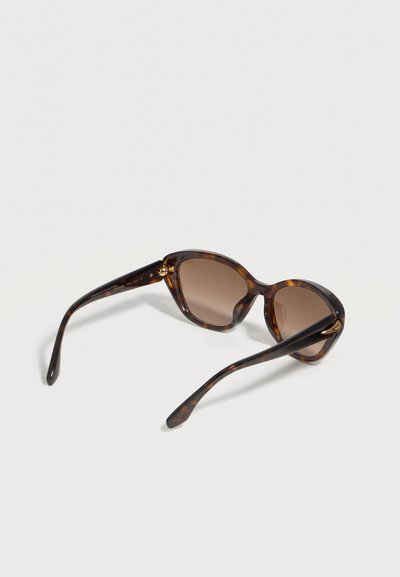 Lunettes de soleil à motif écaille avec des verres carrés arrondis marron, détail de charnière dorée, posées ouvertes sur une surface blanche.