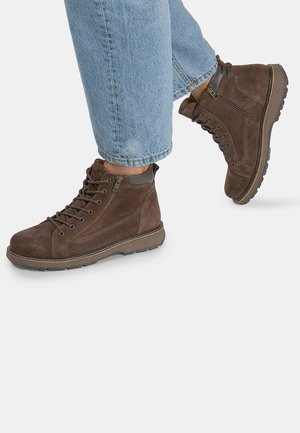 Stivaletti in suede marrone a metà altezza con punta rotonda, lacci frontali, dettaglio con zip laterale e suola in gomma testurizzata. Indossati con jeans in denim chiaro risvoltati.