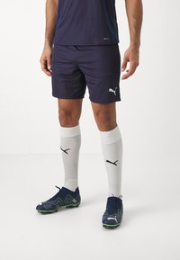 Jūrų sporto šortai su Puma logotipu, derinami su baltais iki kelių siekiančiais kojiniais, turinčiais juodus Puma akcentus, ir mėlynais futbolo batais su žaliais akcentais.
