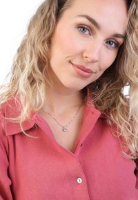 Chemise rose à côtes avec col, portée avec un collier en argent contenant un petit pendentif. Cheveux ondulés et mi-longs.