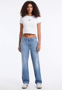 Fehér, rövidített póló fekete csillag grafikával, lazán illeszkedő világoskék farmerrel párosítva. A modell fehér sportcipőt visel fekete díszítéssel.