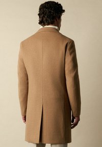 Beiger Wollmantel mit geradem Schnitt, Rückenschlitz und strukturierten Schultern. Glatte Textur und minimalistisches Design.