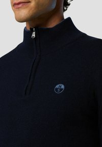 Dunkelblaues Zip-Up Sweatshirt mit hohem Kragen, aus weichem Material, mit einem runden Logo in Hellblau, das "N" und "S" zeigt.