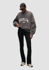 Sudadera gris de gran tamaño con "BERLÍN" en letras blancas, combinada con pantalones ajustados negros. El modelo lleva joyería de plata y gafas de sol.