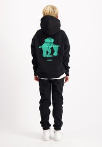 Zwarte hoodie met een levendige groene afbeelding op de achterkant, losse pasvorm, van fleece materiaal. Gecombineerd met zwarte joggingbroek en witte sneakers.