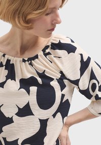 Krémová blůza s námořnickým abstraktním květinovým vzorem, šitým límcem a rukávy do tří čtvrtin. Měkká tkanina s uvolněným střihem.