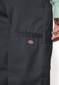 Temno sivi delovni hlače iz trpežne tkanine, z bočnim žepom in pisanim logotipom Dickies. Gladka tekstura, strukturirana zasnova.