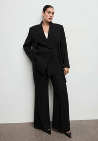 Traje negro a medida con blazer de gran tamaño y pantalones de pierna ancha. Tejido liso con solapas afiladas y bolsillos laterales. Tacones negros puntiagudos.