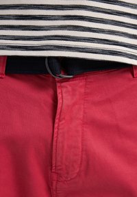 Rode broek met een gladde textuur, voorzien van een rits- en knoopsluiting. Een donkerblauwe geweven riem met een ronde metalen gesp is zichtbaar erboven.
