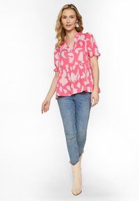 Lyserød bluse med blå og hvide abstrakte mønstre, korte puffærmer og peplum-søm, parret med blå jeans og beige støvler.